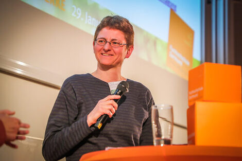 Foto von Professorin Dennert: Frau mit Brille im dunklen Pullover hält ein Mikrofon in der Hand