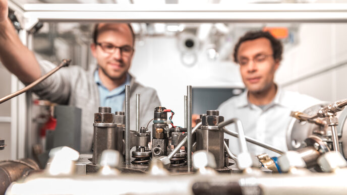 Elements of an engine assembly. Behind it you can see a PhD student and his professor looking at the engine.