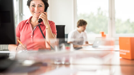 Photo of a female employee sitting at the desk and talking on the phone, smiling at the camera. In the background, a man is working on a laptop.