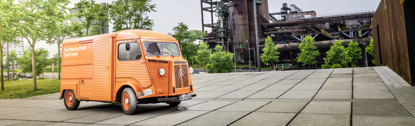 Foto von orangefarbenem Oldtimer-Kastenwagen mit FH-Logo vor Industriedenkmal.___Orange vintage car with FH logo in front of an industrial monument.