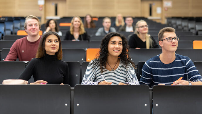 Several students are sitting in a lecture hall. The focus is on a female and a male student sitting next to each other and smiling into the camera.