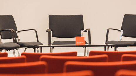 Photo of an empty event room, view over empty rows of chairs to the podium, where there are more chairs and a table with FH cube. __ Empty event room, view over empty rows of chairs to the podium, where there are more chairs and a table with FH cube.