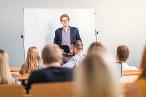 Foto eines Dozenten im Hörsaal, der lachend vor einem Whiteboard steht. Im Vordergrund unscharf erkennbar mehrere sitzende Studierende in Rückansicht.