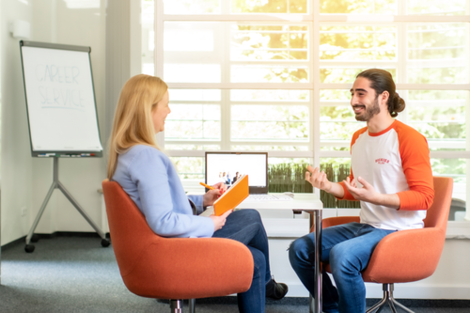 Foto von zwei Personen, die an einem Tisch im Gespräch sind. Die Mitarbeiterin lächelt den jungen Mann, der ihr gegenüber sitz an und macht sich Notizen, Sie ist eher mit dem Rücken zur Kamera gewandt. Der junge Mann strahlt und erzählt mit den Händen. Auf dem Tisch zwischen den beiden sieht man einen Laptop, im Hintergrund links im Bild ist unscharf ein Flipchart zu erkennen.