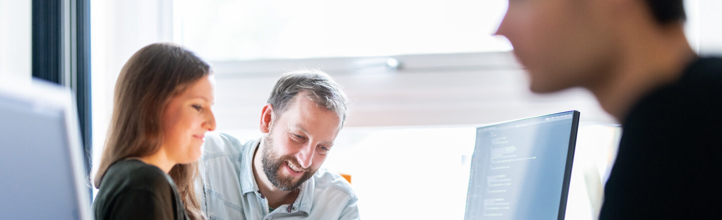 Photo of an employee next to an employee of the Hoschschul-IT, who shows the employee something on the PC. In the foreground, out of focus, another employee at the PC. <br><br>