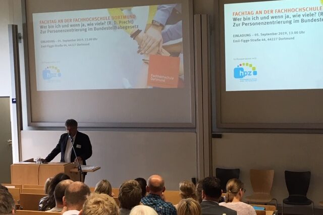 Symposium 2018: A man gives a lecture at a lectern in a lecture hall filled with people. A presentation about the symposium is shown on the screen behind him.