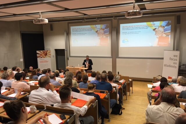 Symposium 2018: A lecture hall full of people listening to a lecturer in front of a screen with a presentation.