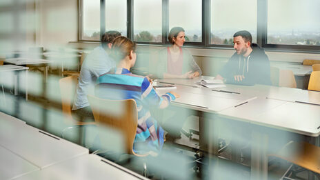 Foto durch eine Fensterscheibe in einen Seminarraum, in dem vier Studierende an einem Tisch sitzen und sich austauschen.