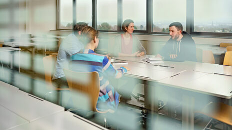 Photo through a window pane into a seminar room where four students are sitting at a table and exchanging ideas.