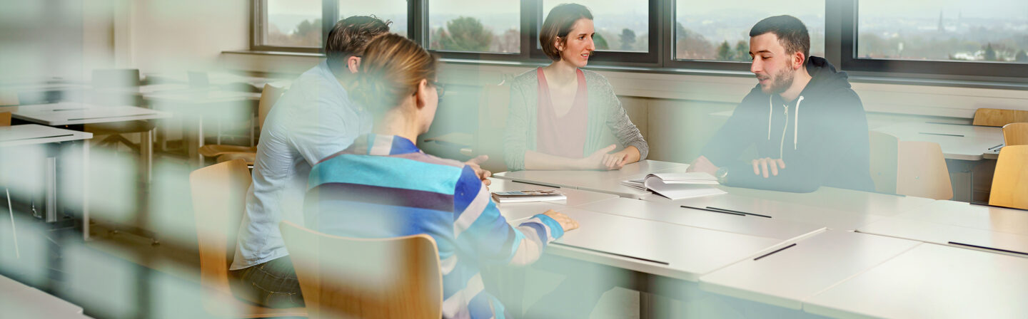 Foto durch eine Fensterscheibe in einen Seminarraum, in dem vier Studierende an einem Tisch sitzen und sich austauschen.