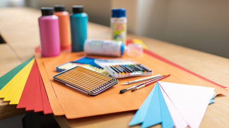 Foto aus der Kreativwerkstatt. Auf einem Tisch stehen und liegen mehrere farbige Pappen, Farbflaschen, Buntstifte, Wachsmalstifte und Pinsel.