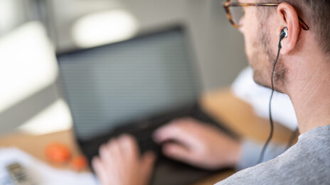 Side photo of a man with in-ear headphones in his ear working on a computer.