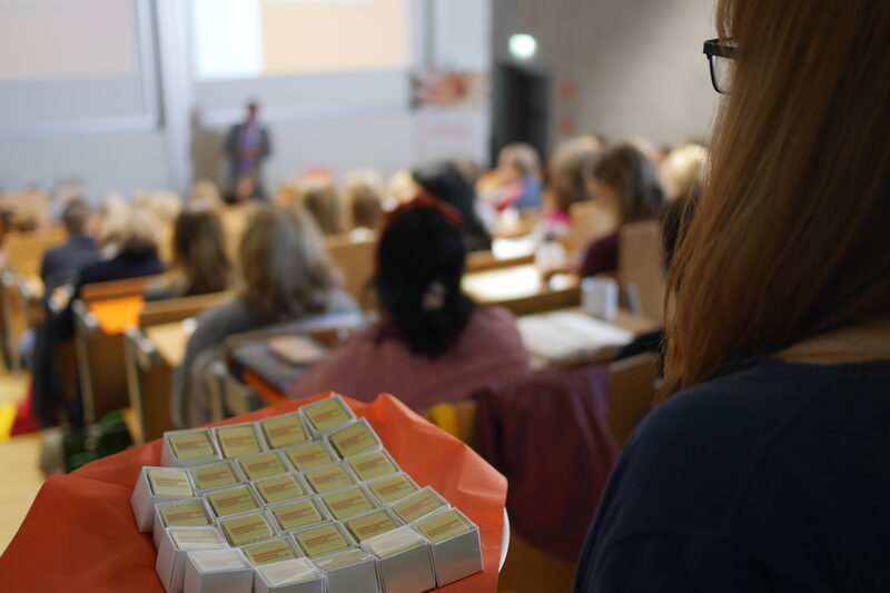 Eine Szene im Hörsaal. Im Vordergrund eine Frau, die einen Teller mit Pralinen mit FH Logo hält. Im Hintergrund sind Personen zu sehen.