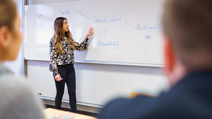 Photo of a young woman standing in front of a whiteboard and explaining a mind map on the subject of social work. Two people listening are out of focus in the foreground.