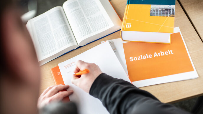 Foto von Jemanden, der etwas mit einem orangefarbenen Stift auf einen Block schreibt. Daneben liegen Bücher und ein Heft mit Aufschrift "soziale Arbeit".