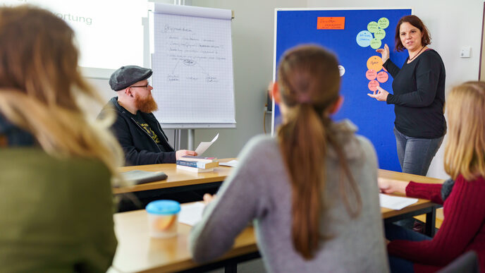 Foto mehrerer Teilnehmer:innen der Arbeitsgruppe Ethnographie, die an Tischen sitzen. Vorne steht eine Frau und erklärt etwas anhand einer Pinnwand mit mehreren bunten Zetteln.