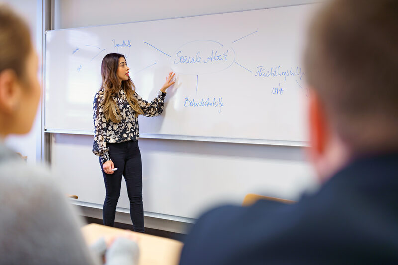 Foto eines jungen Frau, die vor einem Whiteboard steht und daran eine Mindmap zum Thema soziale Arbeit erklärt. Unscharf im Vordergrund zwei Personen, die zuhören.
