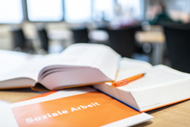 Foto von Büchern, die aufgeschlagen übereinander auf dem Tisch liegen. Davor ein Heft mit der Aufschrift "Soziale Arbeit".