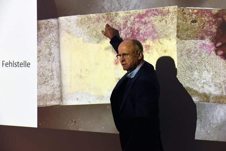 A lecturer stands in front of a projection of a photo. The picture shows a concrete surface with discoloration. The presenter points to a spot in the picture.