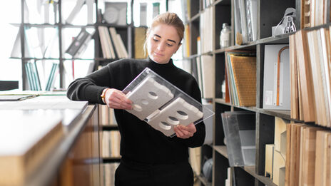 Photo of a woman standing in a room next to shelves and looking at the stone slab she is holding.