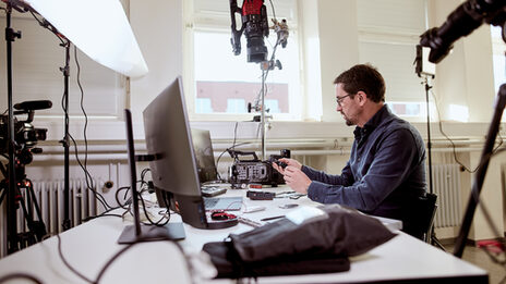 Photo of a lecturer sits at his desk, surrounded by various camera equipment as well as a laptop and monitors, in order to give an online lecture. __ A prof sits at his desk, surrounded by various camera equipment as well as a laptop and monitors, in order to give an online lecture.