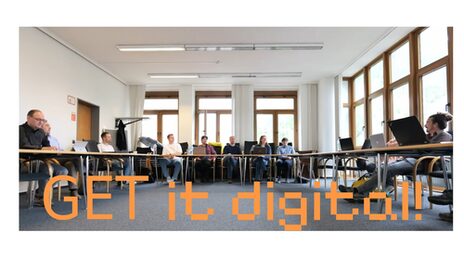 Mehrere Personen nehmen an einer Besprechung in einer offenen U-förmigen Tischanordnung teil. Hinten und rechts im Bild mehrere Fenster. Links hinten die Zugangstür. Unten im Bild in orangener Schrift: „GET it digital!”