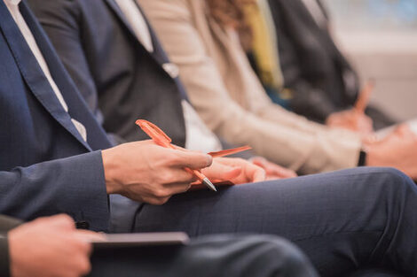 Photo of a row of people sitting at an event. A man holding pen and material in hand. __ Row of people sitting at an event, man holding pen and material in hand.