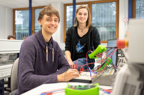 Foto von zwei Studierenden im Labor für Elektronik und Automation. Sie sitzen an bzw. auf einem Tisch mit diversen elektronischen Messgeräten und lächeln in die Kamera.