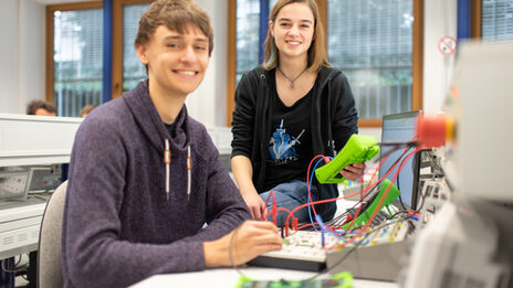 Foto von zwei Studierenden im Labor für Elektronik und Automation. Sie sitzen an bzw. auf einem Tisch mit diversen elektronischen Messgeräten und lächeln in die Kamera.