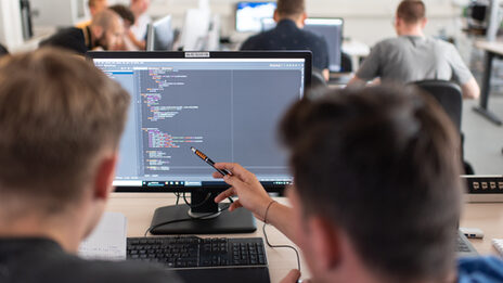 Photo of students in rear view at computer workstations. In the foreground, a student shows something on the screen to the person sitting next to him with a pen.