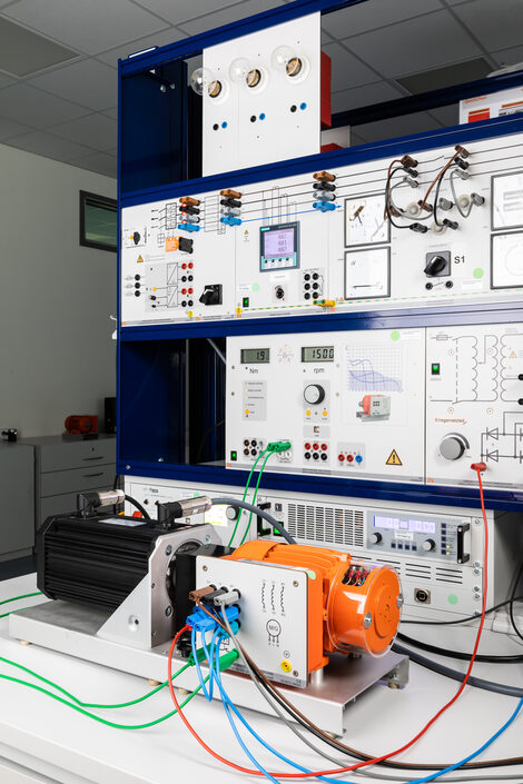 Photo of a test bench for electric motors with power supply and measuring devices, in front of it an orange three-phase motor with coupled load machine on the table.