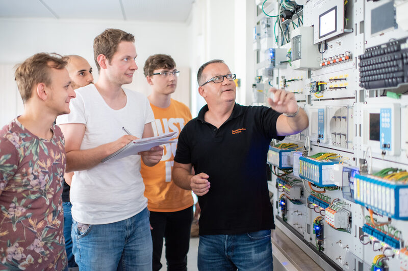 Foto eines Professors mit seinen Studierenden. Sie stehen vor einer Versuchswand der Elektrotechnik, der Professor zeigt auf ein Gerät. Einer der Studierenden macht sich Notizen.