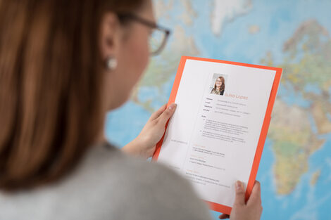 Photo over the shoulder of a woman. She is holding a folder with application documents or a CV in her hands. In the background a world map on the wall. __ Woman holds portfolio with application documents or a résumé in her hands. In the background a world map on the wall.