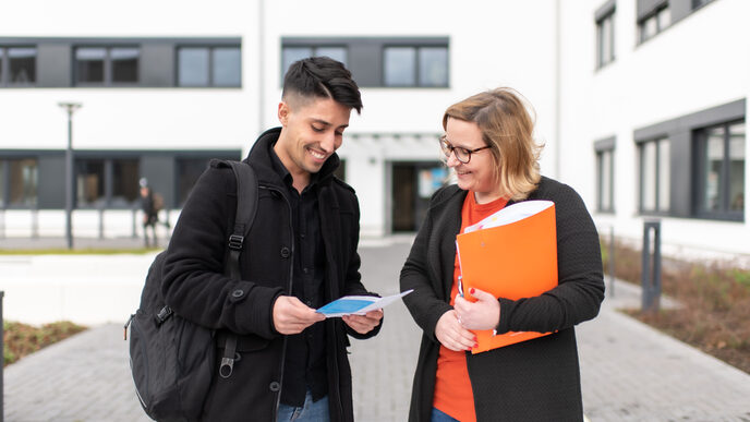 Foto einer Mitarbeiterin und eines Schülers, dir vor einem Campusgebäude stehen. Sie hält eine orangene Mappe unterm Arm, er hat einen Flyer in der Hand. Beide schauen auf den Flyer und lachen.
