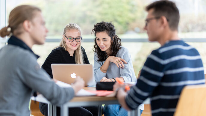 Foto von vier Studierenden an einem Gruppentisch. Im Fokus zwei Studentinnen, die nebeneinander sitzen und auf einen Laptop schauen. Zwei weitere Studenten sind in ein Gespräch vertieft.