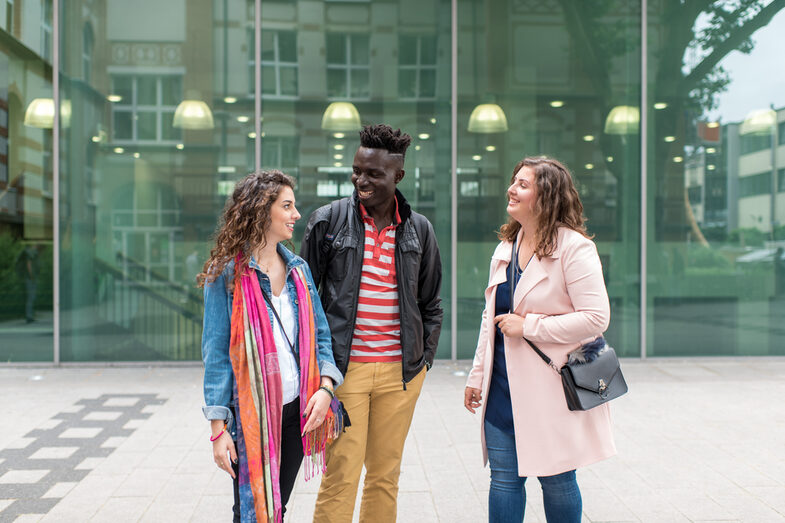 Foto von zwei Studentinnen und einem Studenten, die nebeneinander vor dem Mensagebäude stehen. Sie lachen einander an.__Two female and one male students stand side by side in front of the canteen building, they laugh at each other.