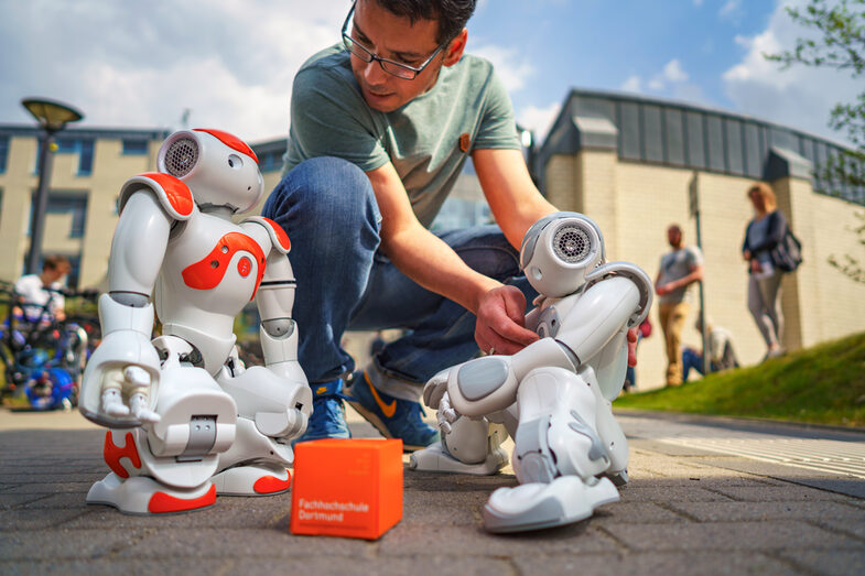 Photo of two NAO robots standing outside Building 42. An employee sets up the robots. A UAS cube lies between the robots.