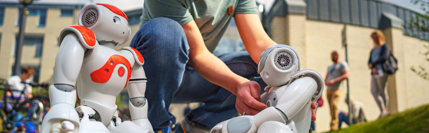 Photo of two NAO robots standing outside Building 42. An employee sets up the robots. A UAS cube lies between the robots.