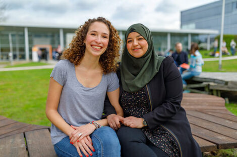 Photo of two female students sitting close together on wooden benches on the campus and smiling at the camera __Two female students sit close together on wooden benches on the campus and smiling at the camera.