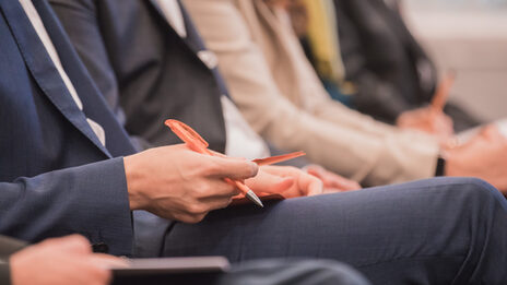 Foto einer Reihe von Menschen, die bei einer Veranstaltung sitzen. Ein Mann hält Kugelschreiber und Material in der Hand. __ Row of people sitting at an event, man holding pen and material in hand.