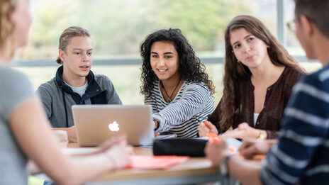 Foto von fünf Studierenden an einem Gruppentisch. Im Fokus eine Studentin und ein Student, die nebeneinander sitzen und gemeinsam auf einen Laptop schauen.