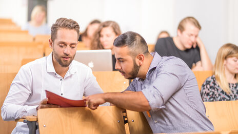 Foto Studierenden im Hörsaal, im Vordergrund zeigt ein Student eeinem anderen Studenten etwas in einer Mappe. Im Hintergrund sitzen weitere Studierende.