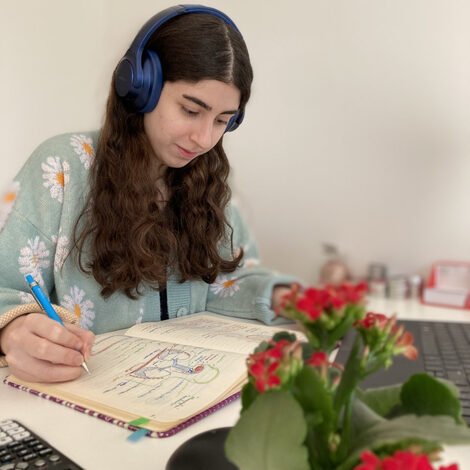 Frau Saleh beim Schreiben in ein Notizbuch vor Ihrem Notebook an einem Tisch Sitzend. Auf dem Kopf trägt Sie Blaue Kopfhörer.<br>Im Vordergrund, unscharf zu sehen, eine Blume.