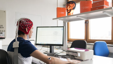 Foto eines Probanden mit EEG Haube und Neurorezeptor an Ellenbogen vor einem Messbildschirm.