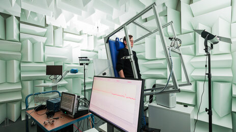 Photo of an experimental set-up for measuring blood pressure using a tilting table. A man is attached to the tilting table in a laboratory, which is almost vertical. Monitors and equipment are set up in front of it.