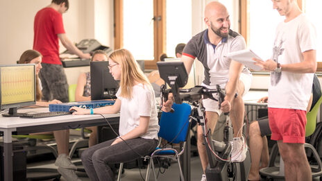 On the right in the photo, a man on a bicycle ergometer and another man next to him taking down values. On the left in the photo, other people at computers.