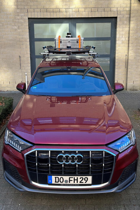 Ein schwarzer Audi Q7 steht vor einem Gebäude. Das Auto hat einen großen Dachgepäckträger, auf dem eine silberne Box mit orangenen Spanngurten und mehrere Sensoren befestigt sind. Das Fahrzeug ist frontal abgebildet und steht auf einem gepflastertem Parkplatz vor einem Gebäude und einer grauen Tür mit mehreren Fenstern.