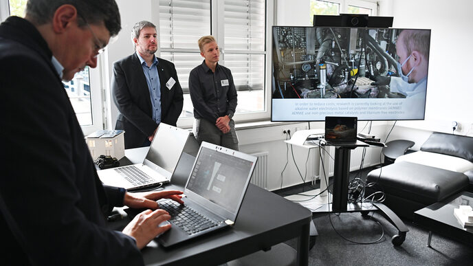 A video is playing on a large screen while a few people can be seen next to it. Another is working on a laptop in the foreground.