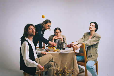 Eine Gruppe von vier männliche gelesenen Personen sitzt um einen runden Tisch auf dem eine Schale voll Obst steht.