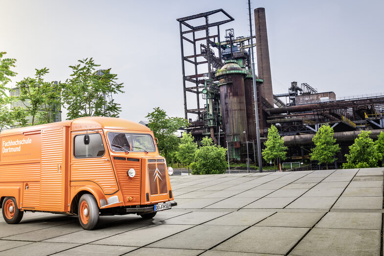 Foto von orangefarbenem Oldtimer-Kastenwagen mit FH-Logo vor Industriedenkmal.___Orange vintage car with FH logo in front of an industrial monument.